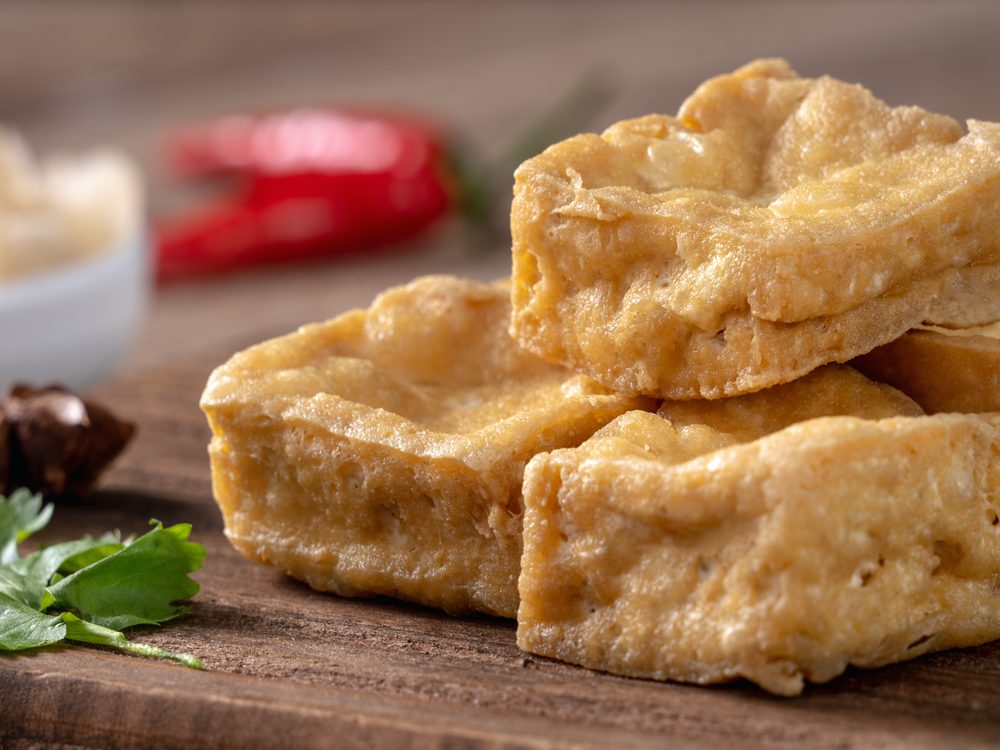 Golden pieces of deep fried stinky tofu stacked on a wooden board with chilli and coriander